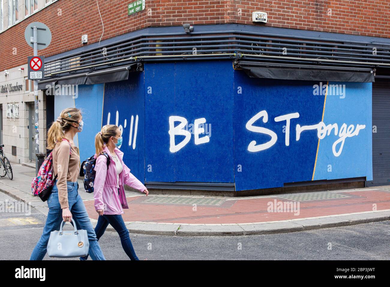 Two girls wearing protective face masks passing by a sign written on the walls of a closed shop - We`ll be strong - during the Covid-19 pandemic. Stock Photo